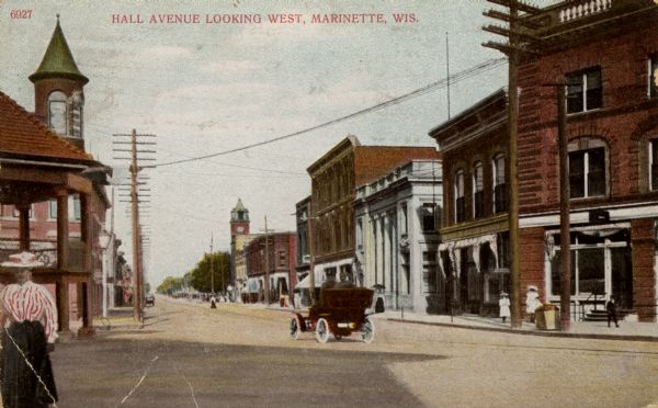 View down avenue. There is a woman in the street in the left foreground, and behind her is a pavilion. An automobile is going down the street, and commercial buildings are along the right. Caption reads: "Hall Avenue looking west, Marinette, Wis."