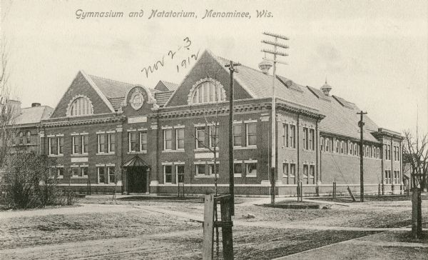 View across unpaved road towards the gymnasium and natatorium. Caption reads: "Gymnasium and Natatorium, Menominee, Wis."
