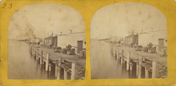 Stereograph of a view along the Fox River. A pier with pilings is along the right, with industrial buildings.