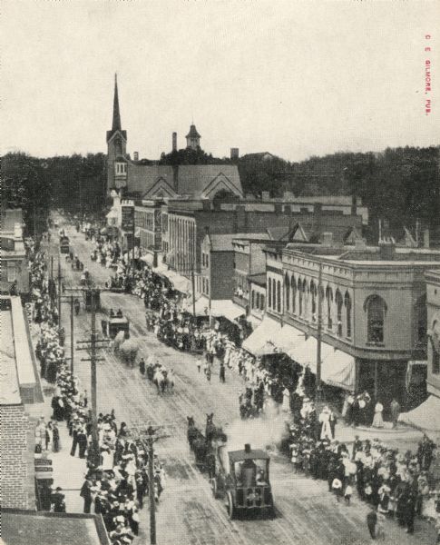 Elevated view of Main Street. Crowds line the sidewalks, and horse drawn vehicles and people are moving down the street.