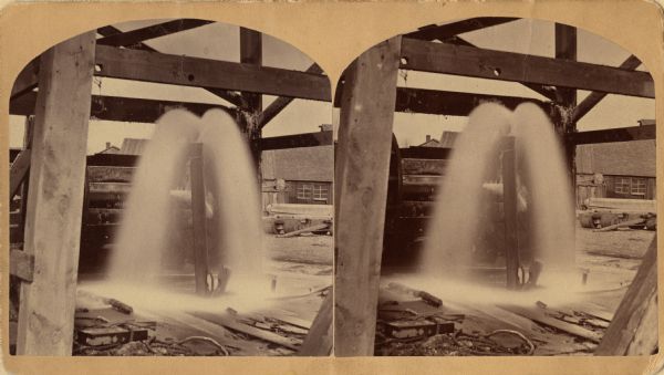 Stereograph of artesian well, apparently not the same one that was located near the intersection of Wisconsin Street and South Minnesota Street (since renamed Wacouta). A wood structure is around the well, and buildings are in the background.