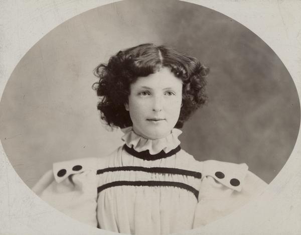Oval portrait of young girl wearing a high-collared dress with puffy sleeves.  She is facing forward, looking slightly to right, with short, wavy hair parted in the middle, wearing a white dress with dark piping across collarbone and beneath ruffed collar, epaulets with two dark buttons, puffed sleeves, looking slightly amused.