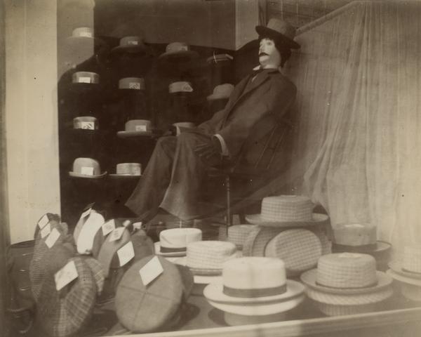 The front window display of a Black River Falls hat shop. Rows of cloth caps, with linen, and straw hats are heaped on the showcase floor. A  mannequin is in the far corner in front of more hats displayed on shelves.