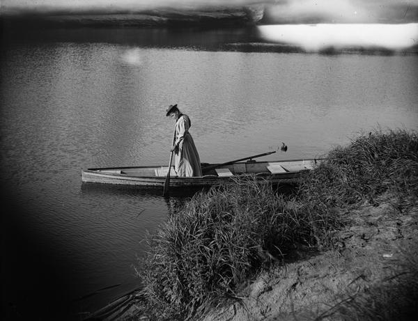 Woman standing in a rowboat near shore, testing the water with an oar.