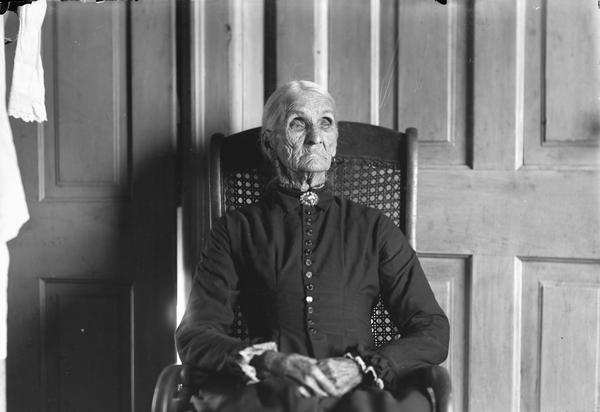 Portrait of a very elderly woman sitting in a wicker wheel chair, in front of two quarter panel doors. She is wearing a dark dress with small buttons to the chin, lace work at sleeves, and a brooch at her neck.  Her hands are crossed in front of her, and she looks slightly to right. Probably Mrs. R.D. Squires.
