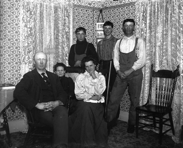 A farm family posing in their living room. Sitting from left to right is John Dietrich (1874-1947), Grace Bowman (1879-1964), and Mrs. Lizzie Macke Dietrich (1880-1933). Standing behind them are Philip (b. 1880), George (b. 1883) and Julius (1885-1952) Nortman. 

Like others in Black River Falls, Dietrich was accused of pro-German sympathies during World War I. He was persecuted by being forced to ride on a log up and down Main Street, etc. On Armistice Day, 1918, a group went to his place in the country, put him in a truck, and made him kiss the United States flag.

Caption from Classified File reads: "Farm family posing in living room. John Diedrich, Grace Bowman and Mrs. Diedrich (Lizzie Macke),--seated. Standing: Philip, George and Julius Nortmann. (Mrs.?) Rozmanoski (owner?) of the Pines Hotel is (Diedrich's) daughter. Like others in this community, Diedrich was accused of pro-German sympathies during the first World War and he was persecuted by being forced to ride a log up and down Main St., etc. On Armistice Day, 1918, a group went to his place out of town, got him in a truck and made him kiss the U.S. flag. (They ? "put the Gerhardt boys on a stone boat").