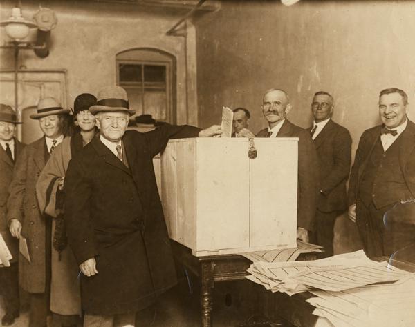Senator Robert M. La Follette, Sr., a third-party presidential candidate, prepares to drop his ballot into the ballot box.  In line behind him are (left to right) his sons, Philip F. La Follette and Robert La Follette, Jr., and Phil's wife, Isabel Bacon La Follette. The man behind the ballot box has been identified as B. Frank Piper.
