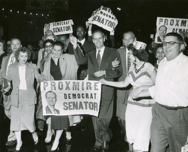 William Proxmire, his wife, Ellen (to Proxmire's right), and his supporters celebrate his victory in the special election to fill the Senate seat vacated upon the death of Senator Joseph R. McCarthy. The supporters carry signs that read "Proxmire Democrat Senator".