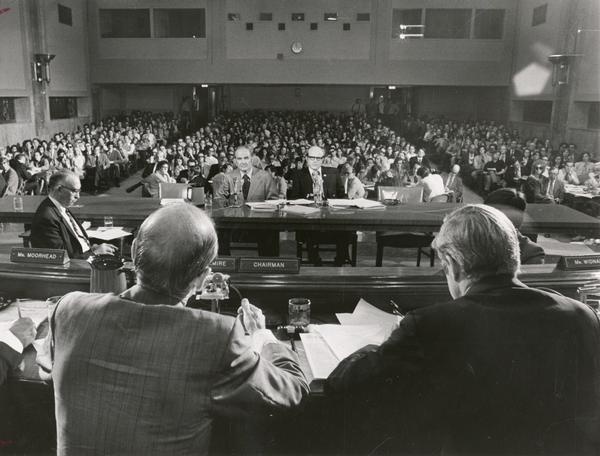 Senator George McGovern of South Dakota testifying before the Joint Economic Committee and a large audience. Senator McGovern was soon to become the Presidential candidate of the Democratic party. Senator William Proxmire, the committee chair, with his back to the camera, is in the left foreground.
