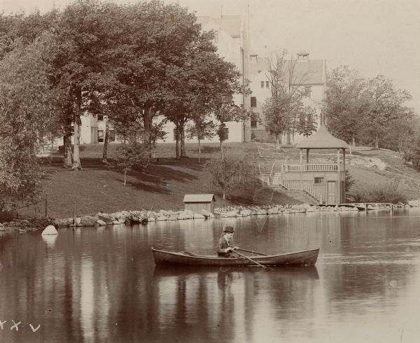 Lake Mendota along the shore of the University of Wisconsin. A boathouse with a roofed pavilion above is on the right. In the foreground is a man rowing a boat in the lake.