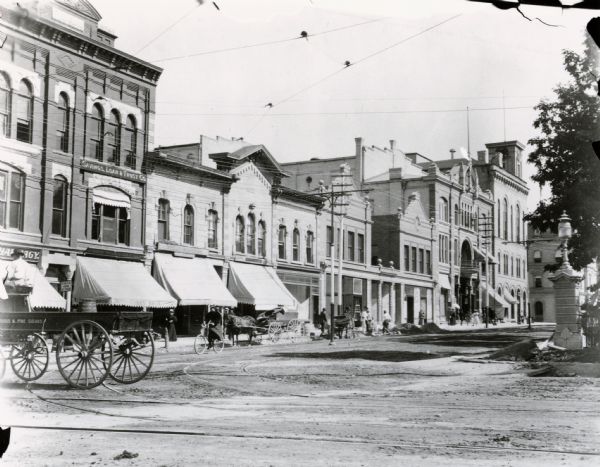 View looking east on West Mifflin Street from its intersection with North Carroll Street on the Capitol Square.  Businesses from right to left: City Hall; Fuller Opera House (with Saul Kasdin fruits & confectionary on one side and Francis Bresee florist on the other side), Wenzel Wirka restaurant, Platz & Essser groceries, Scheler Brothers butchers, Nathaniel Jones photographer (upstairs ?), Halle Steensland's Savings, Loan & Trust Co., Adolph Menges drug store, YMCA (2nd floor) and Kroncke Brothers hardware store.