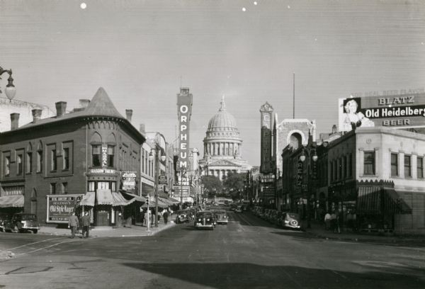 State Street view looking east from Johnson Street, towards the Wisconsin State Capitol. On the corner on the left is Glasgow Tailers, and further up the block is the Orpheum Theatre. On the right corner is the Capitol Tog Shop, Arenz Shoes, and further up the block, the Capitol Theatre.