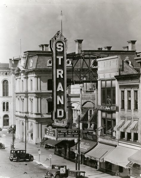 Elevated view of the Strand Theater located at 16 East Mifflin Street near the corner of Wisconsin Avenue. The marquee reads "Babe Ruth and Anna Q. Nilsson, Babe Comes Home." To the left of the theater is an building with an awning that reads: "Collyer Pharmacy." Buildings on the right of the theater have signs that read: "G.R. Kinney Co. Inc. Largest Shoe Retailers," "Hughes Womens Wear," "Buehler Bros. Meats." A street sign reads: "WISCONSIN U S 151."