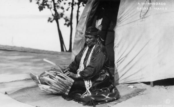 Mary (Stacy) Yellowthunder, (Cowboy Woman) weaving a basket while seated on a blanket. A dwelling and trees can be seen in the background. She is the daughter of Henry Stacy of Wittenburg, Wisconsin. Text at top right reads: "A Winnebago Basket Maker".