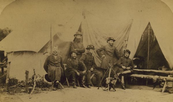 Officers of the Second Wisconsin Infantry posed in front of a tent. They are, from left to right, Quartermaster J.D. Ruggles, Dr. A.J. Ward, Major J.S. Allen (standing), Lt. Colonel Lucius Fairchild, Adjutant C.K. Dean (standing), and Colonel Edgar O'Connor. Fairchild, Dean and O'Connor are clearly wearing the distinctive black hats of the Iron Brigade. The flag of the 2nd can also be seen, unfurled behind them.