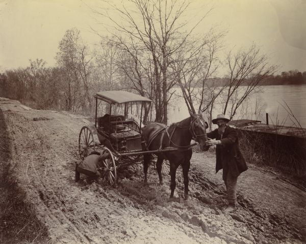 Horse-drawn carriage stuck in a rut on a muddy road near the Ohio River in Floyd County, Indiana. One man is holding the horse while another attempts to dislodge the wheel. This image was entered in a 1898 competition sponsored by the League of American Wheelman to identify the nation's worst roads conditions. The purpose of the competition was to gather evidence of the need for better roads. The donor, Otto Dorner of Milwaukee, was the national chair of the league's road improvement committee.
