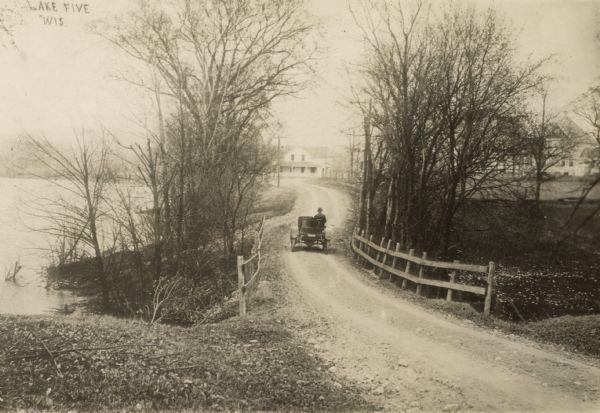 View down dirt road with small bridge along a lake. A man is driving an open automobile towards houses in the background.