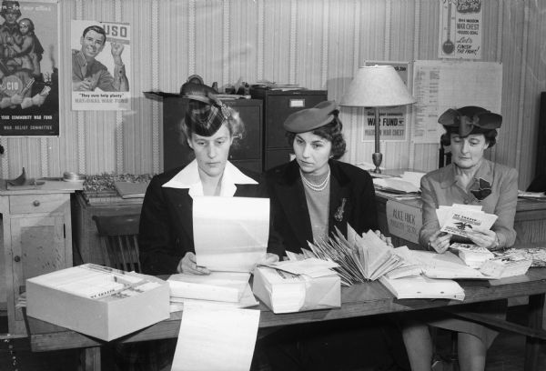 Three female war chest workers seated at a table sort war chest printed material.  The wall in the background is covered with Madison War Chest posters.  Madison War Chest contributes money to the National War fund to support 21 voluntary agencies working for GIs still in service, including USO, and the Allies.