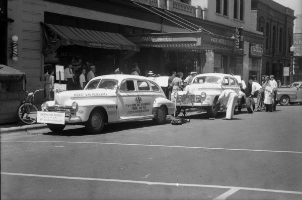 Men doing a periodic rotation of the synthetic and recapped tires on one of the two cars parked in front of the Park Hotel, and participating in the American Automobile Association (AAA), the 25,000 mile "Keep 'Em Rolling" tire safety tour.