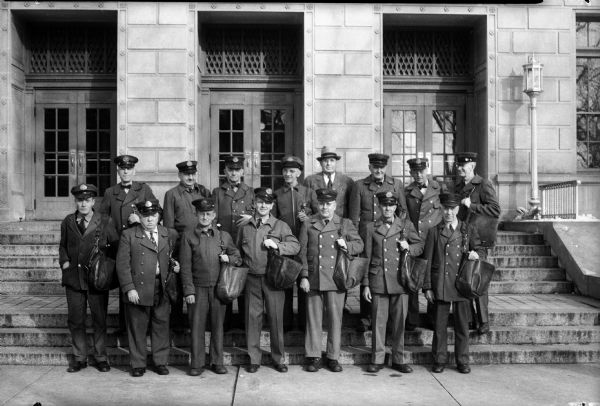 Fifteen Madison U.S. Mail carriers standing on the front steps of the Madison U.S. Post Office. Lower row left to right: Martin T. Digney, Thomas M. Carey, G. Nicholas Mueller, George Flad, Raymond M. Nienaber, Paul W. Kroseman, and Michael Cawley. Upper row left to right: Raymond M. Tierney, Walter S. Fauerbach, Bernhard F. Pahlmeyer, John T. Behrend, Joseph Wirka, Louis F. Nelson, Henry L. Olsen, and Nels E. Anderson.