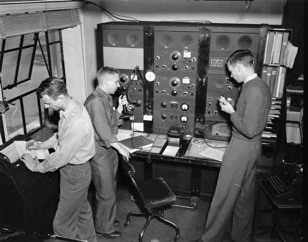 Three men working at the Civil Aeronautics Administration interstate communication station at Truax Field.  From left to right are: Guy J. Blakely checking a weather report; B.W. Dripp, chief aircraft communication in charge of the station; and R.D. Ernst.