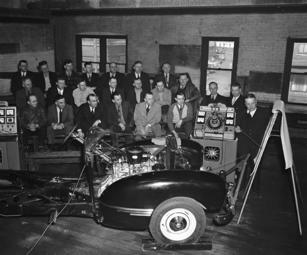Hudson Auto Co., 1134 E. Washington Avenue, holding a "motor school" to teach the use of Sun diagnostic equipment.  Group of men in background, and front end of car in the forefront with diagnostic machine attached.