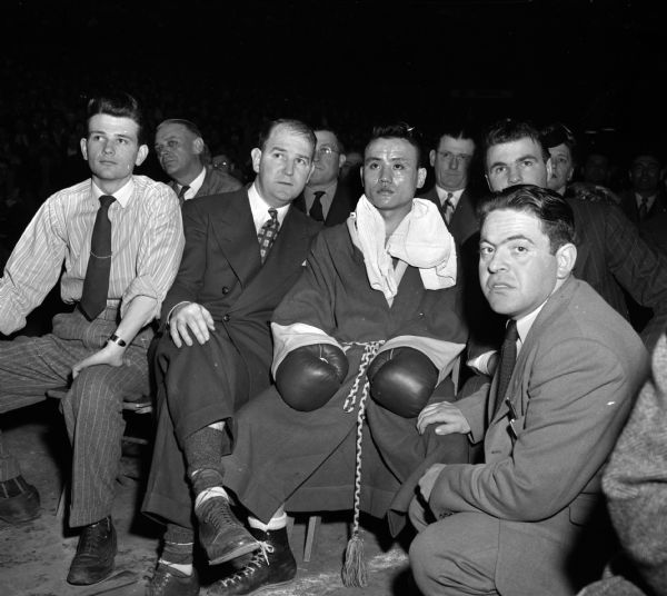 Four University of Wisconsin boxing principals watching a bout at the NCAA National Boxing Tournament at the University of Wisconsin-Madison Field House. From left are John Brandes, a Second; John J. Walsh, Coach; boxer, Dick Miyagwa; and Morrie Holzman, Manager.