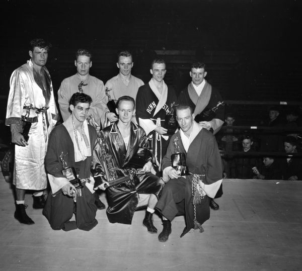 The eight NCAA National Boxing Champions with their trophies. The tournament was held at the University of Wisconsin-Madison Field House. Back row, left to right are: Art Saey, Miami; Laune Erichson, Idaho; Herb Carlson, Idaho; Glen Hawthorne, Penn State; and Gerald Auclair, Syracuse. Front row, left to right are John Lendenski, Wisconsin; Charles Davey, Michigan State; and Cliff Lutz, Wisconsin.