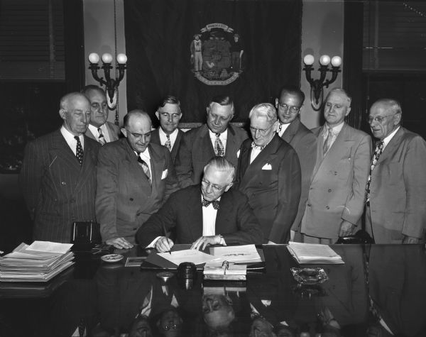 Governor Oscar Rennebohm signing a bill with Voyta J. Wrabetz, Commissioner of the Industrial Commission, and others looking on.