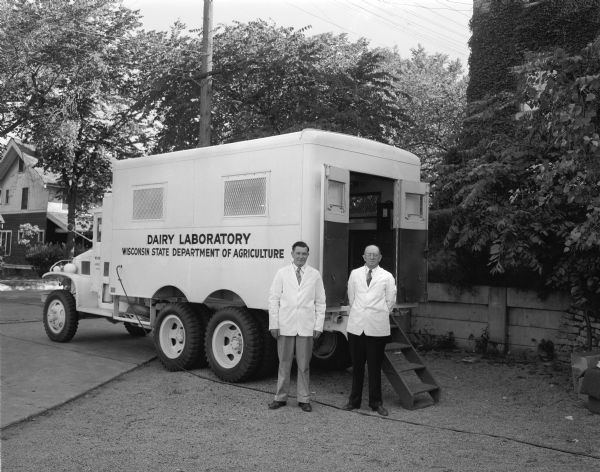 Two men standing at the rear of the Wisconsin Department of Agriculture dairy laboratory truck, which has wooden steps leading up to the open rear doors.