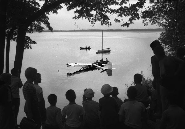 Dozens of boys at Camp Wakanda on the north shore of Lake Mendota watch rescue work on an airplane which has crashed into the lake near shore.  Tom Langlois, 17, 213 North Randall Avenue, was seriously injured when the routine solo training flight crashed into the lake. He was rescued by an unidentified fisherman.