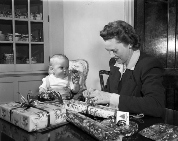 Kenneth Lemmer Jr., sitting in a high chair, is playing with a Christmas tree ornament. His mother Katherine Lemmer is wrapping Christmas presents at a table near a built-in cabinet.