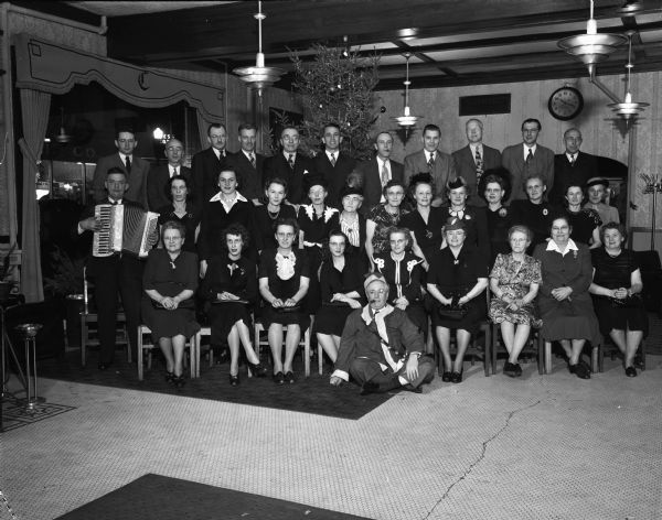 Group portrait of men and women at the Madison Dye Works party.  Included in the portrait on the left is an accordion player. One man sitting on the floor in the front is wearing a Santa Suit.