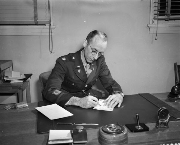 Major Glenn McConagha, commandant of the United States Armed Forces Institute (USAFI) seated at a desk signing a course completion certificate. USAFI provides college level course work to servicemen-students. Seventy colleges and universities are cooperating with USAFI in providing materials and offering credit. USAFI is headquartered at 102 North Hamilton Street.