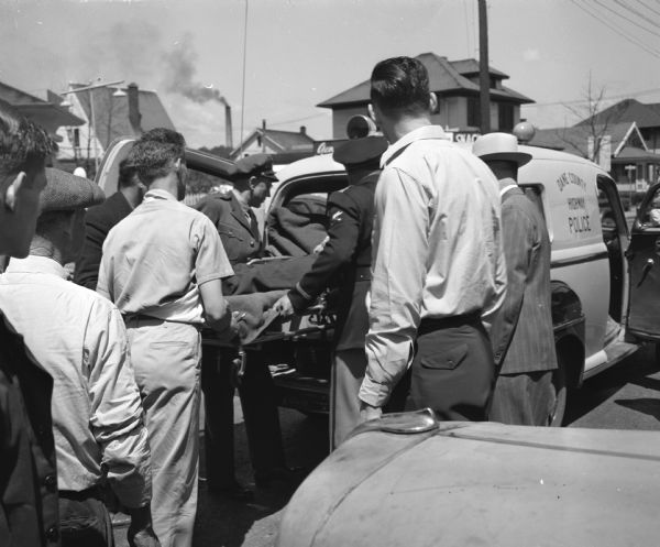 County traffic officers Ray Case (left) and Henry Gerry are placing Elmer C. Evans, with head and face lacerations, in an ambulance to take him to the Jackson clinic. A police ambulance, speeding a 3-year old, David King, seriously injured accident victim to a hospital, crashed into a Padgham Painting Co. truck, driven by Elmer C. Evans, that turned into its path at Park and Regent Streets.