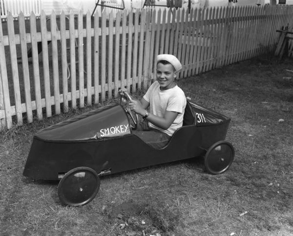 Richard Lerwick, 15, son of Mr. and Mrs. A.W. Lerwick, 1813 Helena Street, shown outdoors near a fence posing in his racing car. He is preparing for the Soap Box Derby on Sunday on East Gorham Street.