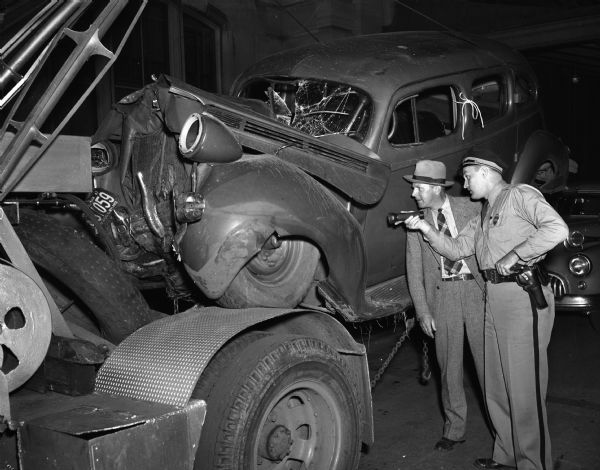 Crashed automobile on tow truck, with traffic officer, Russell Klitzman, and deputy sheriff, Eldon Johnson, viewing the car.