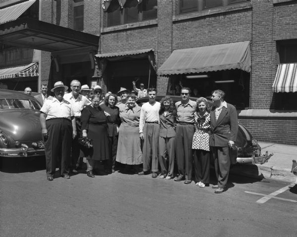 Group portrait of 13 men and women from the Hennie's Shows carnival standing in front of the Wisconsin State Journal building. They had visited Camp Waubeek at Wisconsin Dells to entertain disabled children there. Included are: Jack Norman, Johnnie Johnson, Frank Little, Dean Ardis, the Karns Fat Family, Renee Baron, David Brian, Ed Hayes, Florence Pedajas, and Goodie Holden.