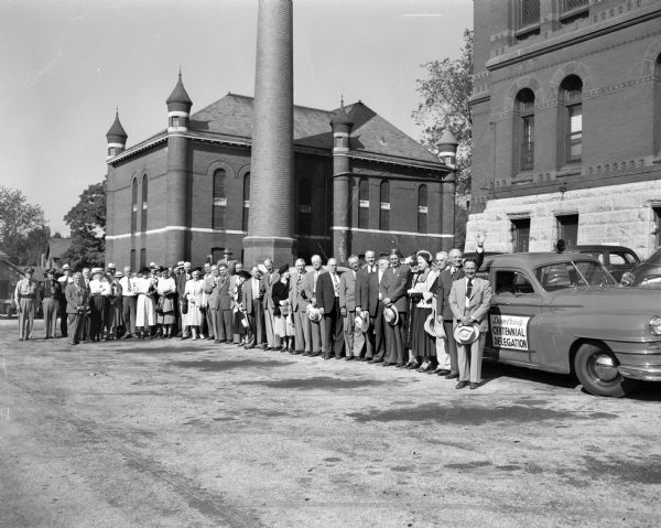 The delegation of state, county, city, university, and business officials is shown as the caravan is about to leave from the Dane County Courthouse, 207 West Main Street, to attend en masse the Madison-Dane County day celebration at the Wisconsin centennial exposition in Milwaukee. The county jail, 219 West Main Street, is also seen in the background. At the right, with his hand raised in greeting, is Governor Oscar Rennebohm. At his right is Chief Justice Marvin B. Rosenberry and the two state officials' wives. Four members of the delegation were injured in a four-car accident while they were traveling in the caravan to Milwaukee.