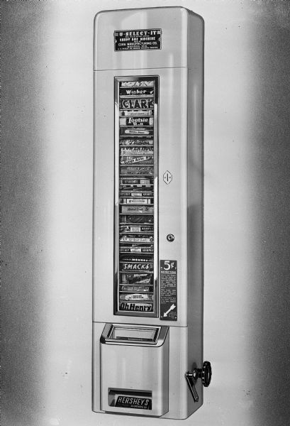 An illustration of a U-Select-It automatic vending machine mounted on a wall and filled with candy bars. Made by the Coan Manufacturing Co., 2070 Helena Street.