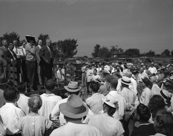 Governor Oscar Rennebohm standing at the podium before a crowd as he auctions off an article at the West Side Business Men's Association auction on Odana Road, site of the proposed new club house.  Standing behind the Governor is Lyall T. Beggs, national commander of the Veterans of Foreign Wars and program speaker at the auction.