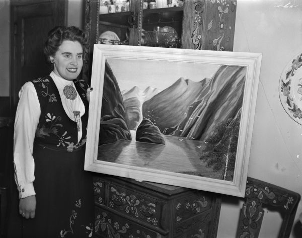 Mrs. Celin Furu is shown in Norwegian costume displaying her oil painting of Trolfjord, as well as Rosemaled furniture. She was a wartime resident of Bergen, Norway when it was bombed 57 times in the month of June, 1940.