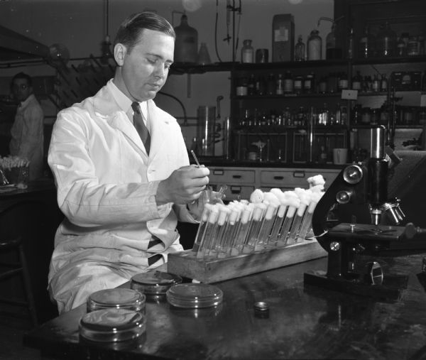 U.S. Army Major Herbert Crecelius of the army chemical corps studying agricultural bacteriology at the University of Wisconsin-Madison. He is in a lab coat with test tubes and a microscope.
