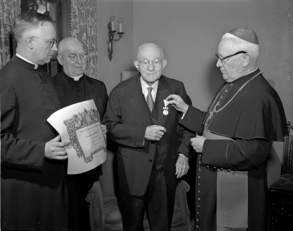Bishop William P. O'Connor presents the medal of Benemerenti to Frank Blied, a member of a pioneer Madison family. The medal, awarded by Pope Pius XII, is in recognition of Blied's work for over half a century in the Catholic Central society. Looking on are the Rev. E.W. Kinney, chancellor of the Madison diocese (left) and the Rev. Joseph H. Gabriels, pastor of Holy Redeemer Catholic church.