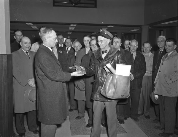 Mailman George A. Perkins receiving a purse containing 25 silver dollars from Marshall Browne, publisher of the East Side News, on the 25th anniversary of his delivering mail on the East Side. Observing the ceremony which took place at the Security State Bank, Atwood Avenue at Winnebago Street, are some 30 East Side business men to whom Perkins has delivered mail during the past 25 years.