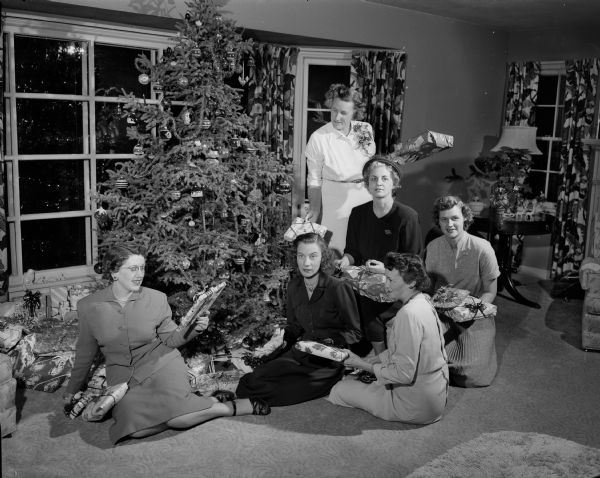 Members of the East Side Women's Club pose around a Christmas tree in Hattie Swan's home at 3738 Atwood Avenue. They are seated with some of the 350 gifts they distributed to the children at Morningside Sanitorium, the Stoughton orphanage and patients at Lake View Sanatorium.  The trio in the foreground include Leona Roman, Jean Scovill and Donna Ellis, president of the East Side Women's Club. The three women in the background are Mrs. Arthur Swan, Minnie Carter, member of the club and also president of the Bell Telephone Co. Auxiliary and Delores Peck.
