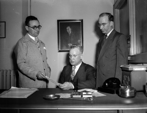 Wisconsin Automotive Trade Association Three men of the Wisconsin Automotive Trade Association sit at and stand by a desk during what is probably a donation check presentation. Organization President, J.P. Gillespie. Organization Executive Secretary, Louis Milan.