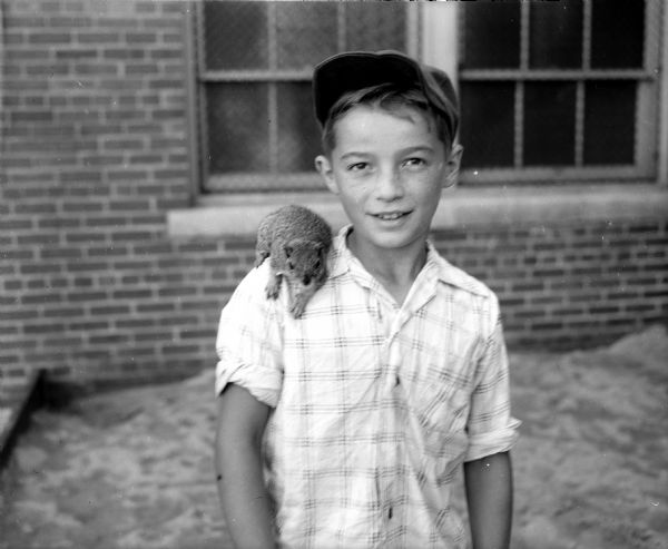 An unidentified boy wearing a baseball cap poses with a squirrel on his shoulder.