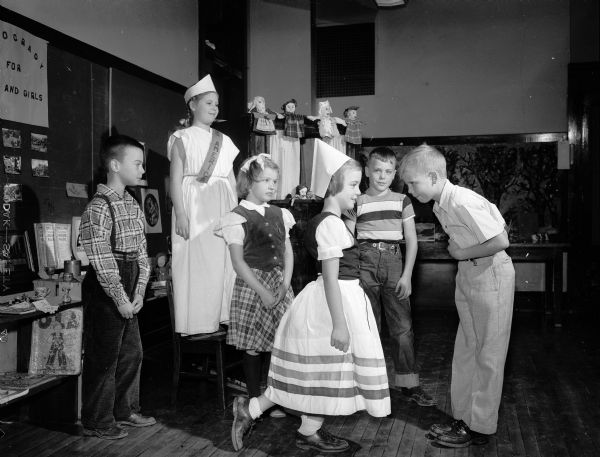 Nakoma School third grade children performing a folk dance as part of a class play, "Democracy for Boys and Girls." The children were assigned to find out where their ancestors came from and what they brought with them to America.  Taking part were, left to right Phillip Goedz, representing Czechoslovakia; Ruth Rauschenberger, Miss America; Alison Keight, Scotland; Barbara Wedell, Norway; David Hofsteen, Holland; and Jerry Morrison, Jr., Italy.  In the background are puppets which the children made and dressed in international costume.