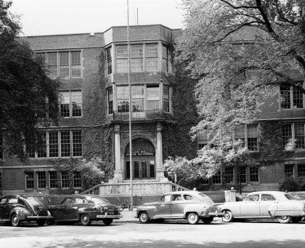 Exterior view of the main entrance to Central High School at Central High School at 214 Wisconsin Avenue. In front of the building are four diagonally parked cars, parking meters, a bicycle, and two shade trees.